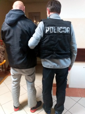 Na zdjęciu widoczny jest korytarz ( w komisariacie policji w Nadarzynie), oraz dwóch stojących plecami mężczyzn. Jeden z mężczyzn po prawej stronie ma na sobie czarną kamizelkę z białym napisem "POLICJA" i trzyma pod rękę drugiego mężczyznę. Mężczyzna ten ubrany jest w czarną kurtkę, jasne spodnie jeansowe i sportowe buty.