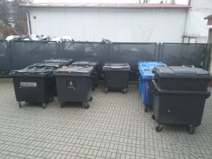 Na zdjęciu widoczne są czarne i niebieskie kontenery na odpady, które są ustawione na terenie parkingu.