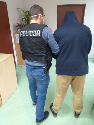 Na zdjęciu widoczny jest policjant, który ma założoną czarną kamizelkę taktyczną z białym napisem policja. Trzyma pod rękę zatrzymanego mężczyznę, który ubrany jest w granatową kurtkę z kapturem nałożonym na głowę, brązowe spodnie i czarne buty.