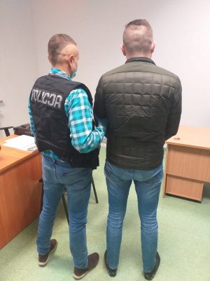 Na zdjęciu widoczny jest policjant, który ma założoną czarną kamizelkę taktyczną z białym napisem policja. Trzyma pod rękę zatrzymanego mężczyznę, który ubrany jest w czarną kurtkę, niebieski spodnie dżinsowe, czarne buty.