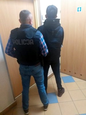 Na zdjęciu widocznych jest dwóch mężczyzn. Znajduję się oni w holu komisariatu w Nadarzynie. Na pierwszym planie widoczny jest policjant ubrany po cywilnemu z czarną kamizelka taktyczną, która posiada biały napis POLICJA znajdujący się na plecach. Policjant trzyma pod rękę zatrzymanego 26-letniego obywatela Słowacji.  Mężczyzna ubrany jest w buty koloru brązowego, ciemne spodnie z ciemną kurtkę z kapturem.