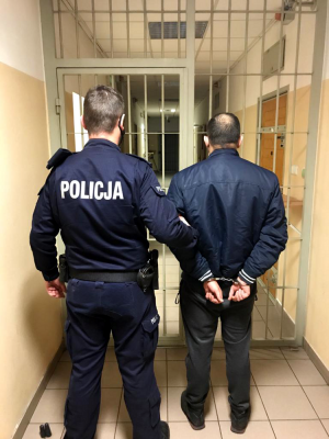 Na zdjęciu widoczne jest pomieszczenie służbowe z krata, przed którym stoi umundurowany policjant i trzyma za rękę mężczyznę zatrzymanego za posiadanie narkotyków. Mężczyzna ubrany jest w ciemne spodnie oraz granatowa kurtkę. Na rękach ma założone kajdanki.