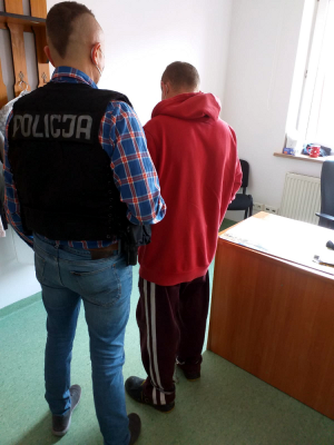 Na zdjęciu widocznych jest dwóch mężczyzn. Jeden z nich ma na sobie czarną kamizelkę taktyczną z białym napisem POLICJA. Drugi mężczyzna zatrzymany 30-latek obrany jest w czerwona bluzę dresową oraz dresowe spodnie bordowe z białymi lampasami na nogawkach. Mężczyzna ma zapięte na nogach kajdanki.