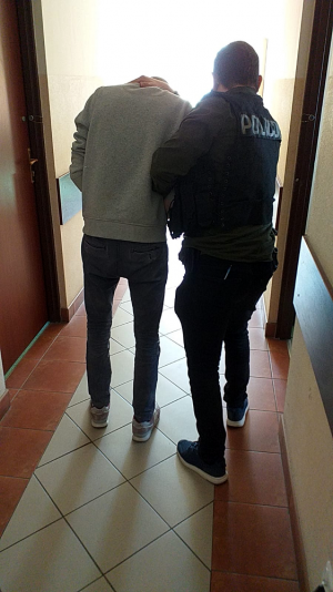 Na zdjęciu policjant ubrany w kamizelkę taktyczną koloru czarnego z białym napisem POLICJA, prowadzi zatrzymanego mężczyznę, który ubrany jest w szara bluzę dresową oraz spodnie jeansowe.