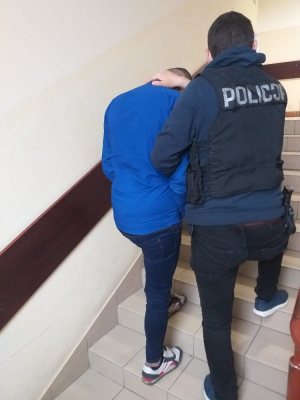 Na zdjęciu widoczny jest policjant ( w kamizelce taktycznej z napisem Policja), który prowadzi zatrzymanego mężczyznę po schodach. Mężczyzna ubrany w niebieską bluzę.