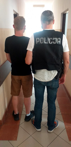 Na zdjęciu widoczny jest policjant w kamizelce taktycznej koloru czarnego z białym napisem  POLICJA, który prowadzi młodego mężczyznę ubranego w krótkie spodenki koloru brązowego i czarną koszulkę z krótkim rękawem.