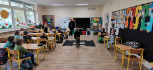 Na zdjęciu policjanci z Komendy Powiatowej Policji w Pruszkowie wspólnie z przedstawicielką Warszawskiej Kolei Dojazdowej przeprowadzają zajęcia o bezpieczeństwie przy przejazdach kolejowych i stacjach w klasach 0-3 w Szkole Podstawowej w Otrębusach.