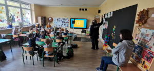 Na zdjęciu policjanci z Komendy Powiatowej Policji w Pruszkowie wspólnie z przedstawicielką Warszawskiej Kolei Dojazdowej przeprowadzają zajęcia o bezpieczeństwie przy przejazdach kolejowych i stacjach w klasach 0-3 w Szkole Podstawowej w Otrębusach.