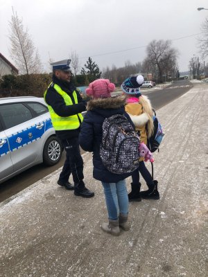 Na zdjęciu widoczny jest umundurowany policjant z Wydziału Ruchu Drogowego Komendy Stołecznej Policji, który rozdaje odblaski dwóm dziewczynkom w drodze do szkoły.