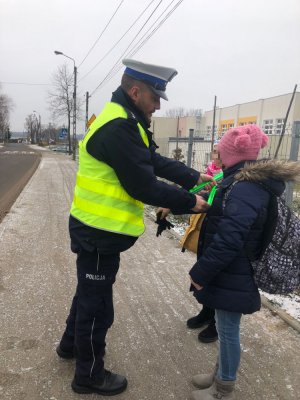 Na zdjęciu widoczny jest policjant, który przymocowuje elementy odblaskowe dziewczynkom w drodze do szkoły.