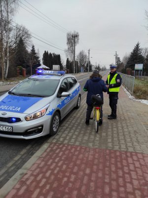 Na zdjęciu widoczny jest radiowóz oraz policjant, który wręcza rowerzystce elementy odblaskowe.