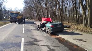 Na zdjęciu widoczne jest miejsce wypadku drogowego ze skutkiem śmiertelnym. Widać pojazd marki peugeot koloru zielonego, który nie posiada przodu, przez tym pojazdem widoczny jest czerwony parawan a po lewej stronie widoczna jest czarno żółta ciężarówka marki scania.