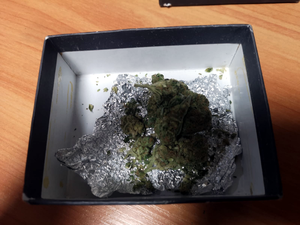 Na zdjęciu widoczne jest kartonowe pudełko w kolorze biało-czarnym, w środku znajduje się susz roślinny koloru zielonego -marihuana.