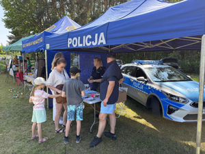 stoisko policyjne, pod niebieskim namiotem z napisem białym policja, przed stoiskiem stojący: dwie kobiety, mężczyzna, dwoje dzieci. Przed nimi umundurowana policjantka