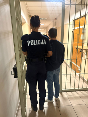 umundurowana policjantka stojąca tyłem z zatrzymanym mężczyzną w wejściu zakratowanym